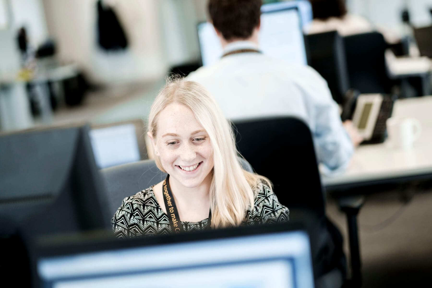 Person with blonde hair using a computer with people working in the background (Skills Hub Help)