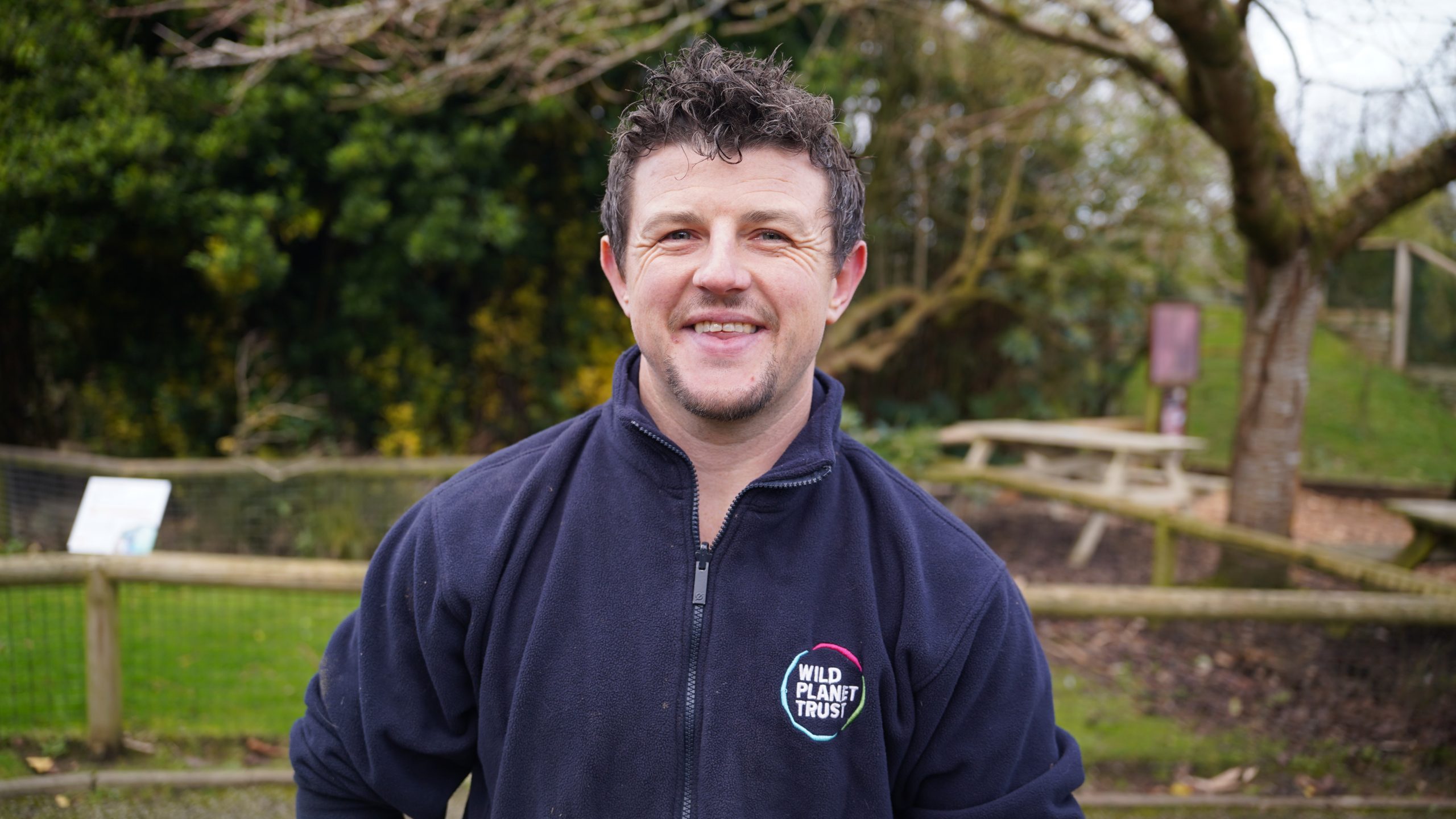 Person smiling with trees and bushes behind them