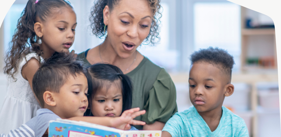 A group of children and an adult are reading books together. The setting appears to be a classroom or library, with shelves of books and educational materials in the background.