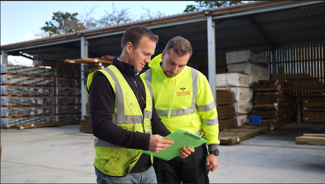 Person in a hi-vis holding a clipboard with another person in a hi-vis looking over his shoulder in a works yard
