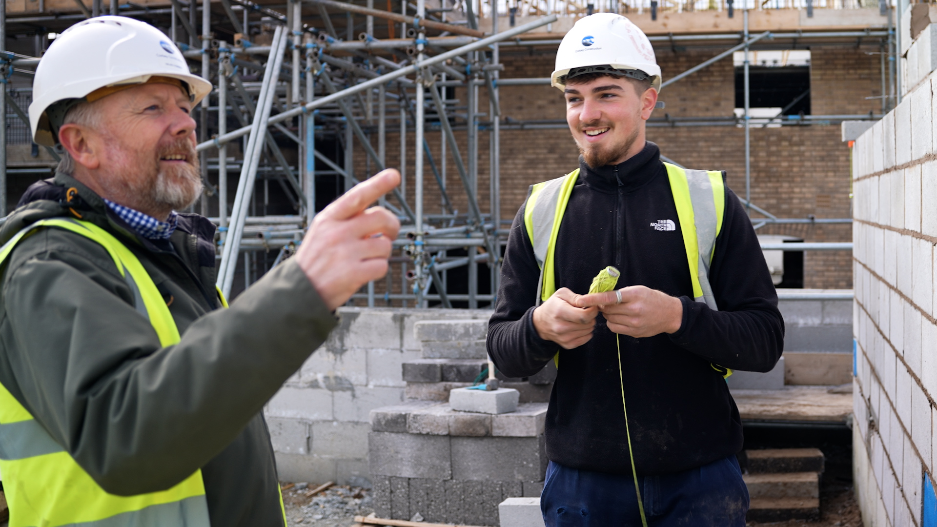 older man in hi-vis and hard hat pointing at something in a building site with a younger man wearing a hard hat and hi-vis smiling
